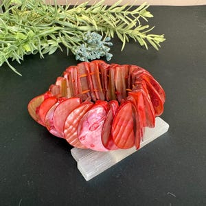 Puede incluir: Un brazalete de concha rojo y rosa. El brazalete está hecho de conchas pulidas en forma de abanico ensartadas. Las conchas varían en tonos de rojo y rosa, algunas muestran cualidades iridiscentes. El brazalete descansa sobre un bloque rectangular blanco.