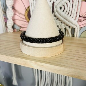May include: A black beaded bracelet displayed on a wooden jewelry stand. The bracelet is a dark, solid color, and the beads appear to be small and uniform. The stand is light-colored wood, and the background includes a floral print and macrame.