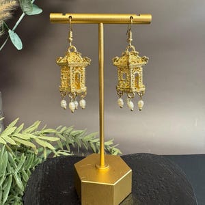 Puede incluir: Pendientes dorados con forma de pagodas ornamentadas, cada uno con perlas blancas colgantes. Los pendientes se exhiben en un soporte dorado sobre un fondo neutro, mostrando su intrincado diseño y estilo elegante.