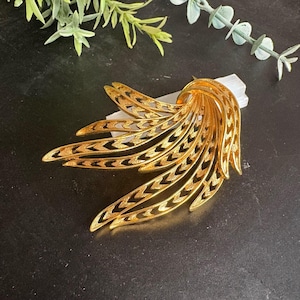May include: A gold-toned vintage brooch with a stylized feather or wing design. The brooch features a series of curved, openwork elements with a chevron pattern, creating a layered effect. The piece is likely made of metal and has a polished finish.