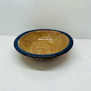 May include: A small, handmade ceramic bowl with a circular design in the center. The bowl has a tan interior and a dark blue rim. The exterior is a light tan color. This bowl is suitable for serving small portions of food.