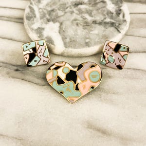 May include: A set of three enamel pins: two square and one heart-shaped. The pins feature a geometric design with black, pink, light blue, and gold accents. The heart pin is in the center, flanked by the square pins.