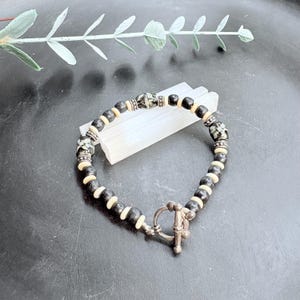May include: A beaded bracelet featuring black and cream-colored beads, interspersed with decorative patterned beads. The bracelet has a silver-toned toggle clasp. The jewelry is displayed on a white rectangular object, with a dark background and a sprig of greenery.