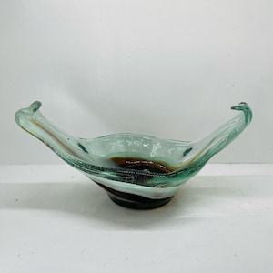 May include: A decorative glass bowl with a unique shape and color. The bowl has a light green and brown color scheme with a wavy design. The bowl has two handles that extend upwards. The bowl is likely used for decorative purposes.