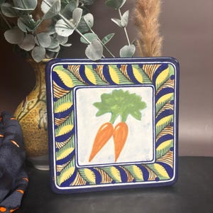 May include: Square ceramic tile with a hand-painted design of two orange carrots with green tops on a white background. The tile has a blue border with yellow and green leaf-like patterns. Decorative kitchenware.