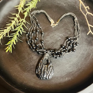 May include: A black necklace with a teardrop pendant. The pendant has a textured surface and a hole near the top. The necklace features multiple strands of black beads and a clasp. The necklace is displayed on a dark surface with greenery.