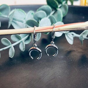 Könnte beinhalten: Ein Paar silberfarbene Halbmond-Ohrringe. Die Ohrringe hängen an einem Holzstab, mit grünen Blättern im Hintergrund. Die Ohrringe haben ein schlichtes, elegantes Design.