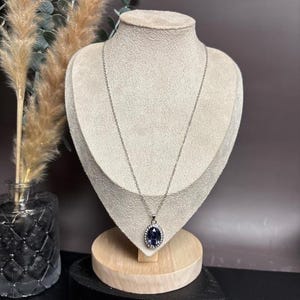 May include: A silver-toned necklace with a large, oval, dark blue pendant. The pendant is surrounded by a halo of small, clear crystals. The necklace is displayed on a mannequin, showcasing its length and design. The background includes decorative elements.