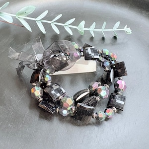 Op de afbeelding: Een zwarte armband met vierkante en facetgeslepen kralen. De armband heeft iriserende kralen en is versierd met een zwart lint. De sieraden worden gepresenteerd op een donkere ondergrond met een takje groen.