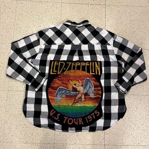 Może przedstawiać: Czarno-biała koszula w kratę z nadrukiem Led Zeppelin na plecach. Grafika przedstawia anioła ze skrzydłami oraz napis "LED ZEPPELIN U.S. TOUR 1975".