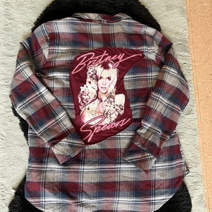 May include: A red and blue plaid flannel shirt with a large patch on the back featuring a photo of Britney Spears with the text "Britney Spears" in pink neon lettering.