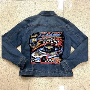 Pode incluir: Jaqueta jeans azul com um grande patch colorido nas costas. O patch apresenta um design de carro de corrida com o texto "Van Halen Racing" e o número "84". A jaqueta tem um design clássico com botões.