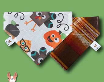 Op de afbeelding: Twee hondenbandanas, een met een witte achtergrond en een Thanksgiving-thema print met kalkoenen, pompoenen en brillen, en de andere met een bruin en oranje ruitpatroon. Beide bandanas hebben een wit label met de tekst "Happy Hounds".