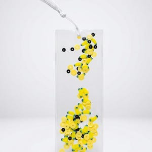 May include: A rectangular clear bookmark containing small yellow banana-shaped charms and black circular sequins suspended inside. A white cord is attached to the top of the bookmark, ending in a white tassel that hangs freely from the top edge.