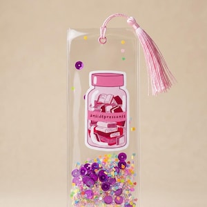 May include: A clear rectangular bookmark featuring a pink tassel and a central graphic of a jar filled with books, labeled with the text 'antidepressants'. The bottom section contains loose purple sequins and multicolored confetti-like beads.