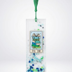 May include: A rectangular resin bookmark featuring a central tarot-style card illustration of a stack of books with the text THE TBR. The clear resin is filled with blue, green, and silver sequins and glitter, finished with a green fabric tassel.