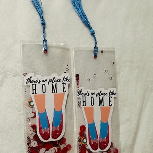 May include: Two clear plastic bookmarks with blue tassels. Each bookmark features a red glitter shoe with a blue sock and the text "there's no place like HOME". The bookmarks are filled with red, white, and rainbow glitter.