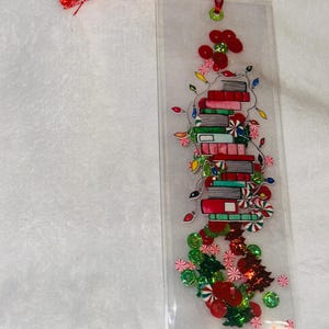 May include: A clear plastic bookmark with a red tassel. The bookmark features a stack of books decorated with Christmas lights, candy canes, and green and red sequins.