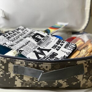 May include: A camouflage-patterned lunch bag is open, revealing a variety of items. Inside, a newspaper-style fabric item with "Harry Potter" text is visible, along with a clear container of snacks and other colorful items.