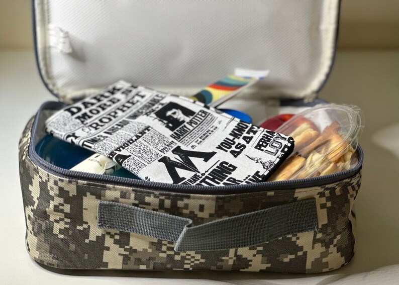 May include: A camouflage-patterned lunch bag is open, revealing a variety of items. Inside, a newspaper-style fabric item with "Harry Potter" text is visible, along with a clear container of snacks and other colorful items.