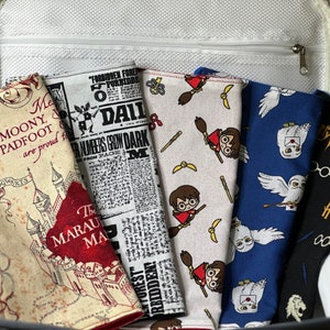 May include: A collection of fabric items with various Harry Potter-themed patterns, including Hogwarts, Daily Prophet newspaper clippings, and characters like Harry Potter and Hedwig. The items are in a gray and white container.