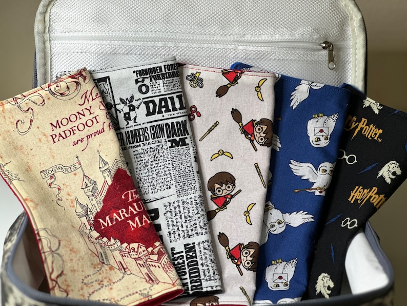 May include: A collection of fabric items with various Harry Potter-themed patterns, including Hogwarts, Daily Prophet newspaper clippings, and characters like Harry Potter and Hedwig. The items are in a gray and white container.