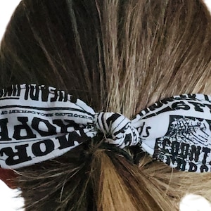 May include: A white and black fabric hair bow tied in a knot, with the bow ends featuring a newspaper print design. The hair accessory is worn in a ponytail, with the bow positioned at the base of the hair.