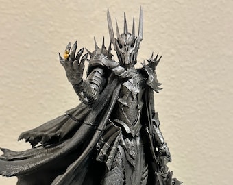 May include: A black and silver figurine of the Lord of the Rings character, The Witch King of Angmar, holding the One Ring. The figurine is standing on a base with skulls.