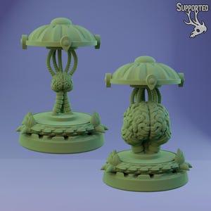 Op de afbeelding: Twee lichtgroene, futuristisch ogende sculpturen. Elke sculptuur heeft een hersenvormig middelpunt, verbonden met een schijfvormige bovenkant met gebogen buizen. De basis heeft een cirkelvormig ontwerp met bloemaccenten. Op een paarse achtergrond.