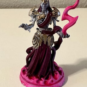 Op de afbeelding: Een geschilderd miniatuurbeeldje van een skeletfiguur dat een gouden en paarse robe draagt en een roze bliksemstraal vasthoudt. Het beeldje staat op een roze voet.