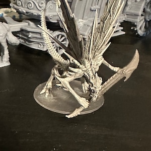 Puede incluir: Una criatura alada esquelética gris con una cola larga y delgada y una hoja afilada y puntiaguda. La criatura está en una postura de ataque dinámica, con las alas extendidas y la hoja levantada en alto.