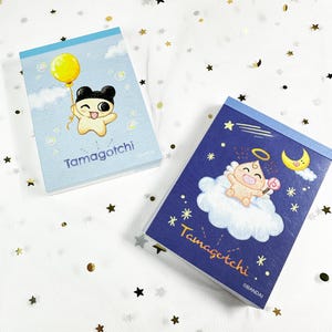 May include: Two Tamagotchi-themed notepads. One notepad has a light blue cover with a cartoon character holding a yellow balloon. The other notepad has a dark blue cover with a cartoon character on a cloud. The text "Tamagotchi" is visible on both.