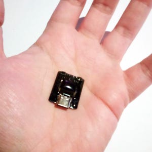 May include: A small, black electronic component with a USB port is held in a person's hand. The component has a rectangular shape with a glossy finish and a silver-colored USB connector at the bottom.