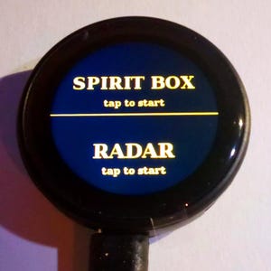 May include: A circular black electronic device with a dark blue screen displaying yellow text. The screen shows the words SPIRIT BOX with tap to start below it, a horizontal yellow line, and the word RADAR with tap to start below it.