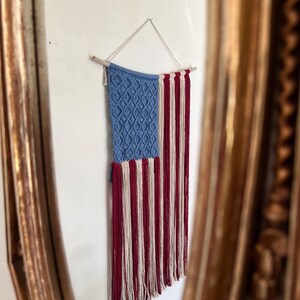 Puede incluir: Un tapiz de macramé que representa la bandera estadounidense. La esquina superior izquierda es azul con un patrón de diamantes, y el resto presenta rayas rojas y blancas. Está suspendido de una varilla de madera con un cordón blanco.