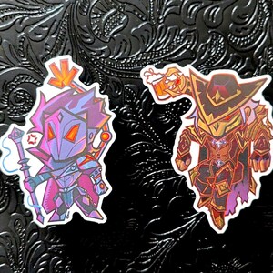 May include: Two stickers featuring stylized fantasy characters. One is a purple and blue mage with a staff, the other a red and gold armored figure with a beer stein. Both have a cartoonish design.