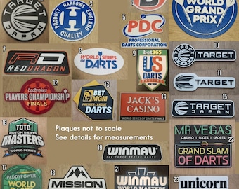 May include: A collection of dart-related logos and plaques, featuring brands like Target, Winmau, and PDC. The plaques vary in shape and color, with text such as "World Grand Prix" and "Dutch Darts Masters".