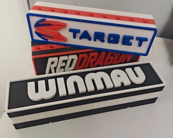 May include: Three rectangular dartboard accessories are stacked. The top one is white with red and blue accents and the word "TARGET" in blue. The middle one is black with red text that reads "RED DRAGON". The bottom one is black with white text that reads "WINMAU".