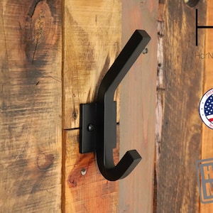 May include: Black metal hook mounted on a wooden wall. The hook is shaped like a letter 'Y' and has a square base. The hook is labeled "HarNa Design" and "Handmade".