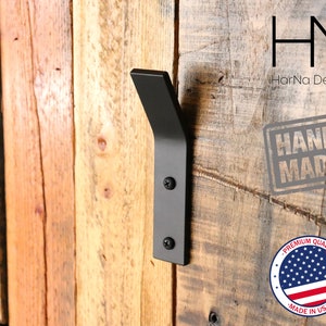 May include: Black metal wall hook with a curved design, mounted on a wooden wall. The hook is labeled "HN HarNa Design" and "HAND MADE".