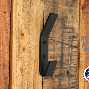 May include: Black metal wall hook with a unique curved design. The hook is mounted on a wooden wall. The text "HarNa Design" and "HANDMADE" is visible in the image.