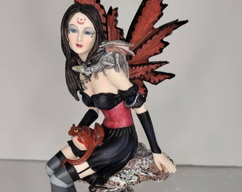 May include: A dark-haired fairy figurine with red wings sits on a rock formation. She is holding a small red dragon and a silver dragon is perched on her shoulder. The base of the figurine is decorated with broken eggs and small dragons.