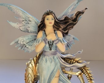 May include: A blue fairy figurine with wings and long brown hair, standing on a rock base with a golden skeletal dragon. The fairy is wearing a blue dress and a crown.