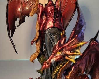 May include: A fantasy figurine of a winged woman in a red bodice and long black skirt, with a dragon at her feet. The woman has long blonde hair and pointed ears. The dragon is red and gold, with detailed scales and wings.