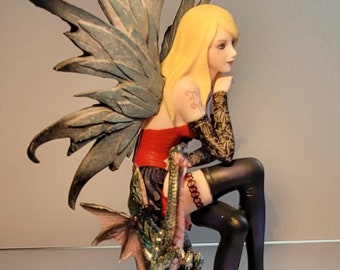 May include: A figurine of a fairy with blue wings sitting on a rock formation with a small green dragon. The fairy is wearing a red and black dress and black stockings.