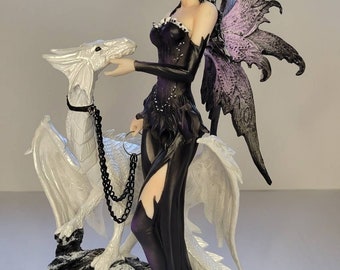 May include: A figurine of a fairy in a black dress with a white dragon. The fairy has black hair and purple and black wings. The dragon is white with a black chain leash. The base is a rocky, gray surface.