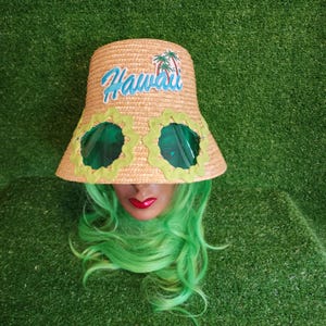 May include: A tan straw bucket hat featuring the word Hawaii in blue script text with a small palm tree graphic above it. The hat includes two circular green tinted lenses framed by a wavy light green fabric trim attached to the front brim.
