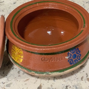 May include: A terracotta cooking pot with a green rim and painted floral designs in yellow, blue, and green. The pot has a ClayStock logo on the side.
