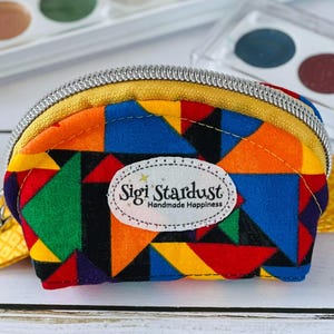 May include: A colorful, geometric patterned coin purse with a silver zipper and a yellow trim. The purse has a label that reads "Sigi Stardust Handmade Happiness." Colored pencils and makeup palettes are in the background.