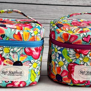 May include: Two cylindrical fabric bags with handles, featuring a vibrant floral pattern in shades of pink, orange, yellow, and white on a turquoise background. Each bag has a zipper and a white rectangular label reading Sigi Stardust.